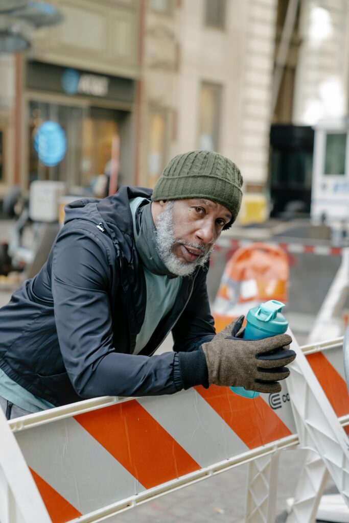 An adult man in casual wear leans on a barricade with a drink in a cityscape.